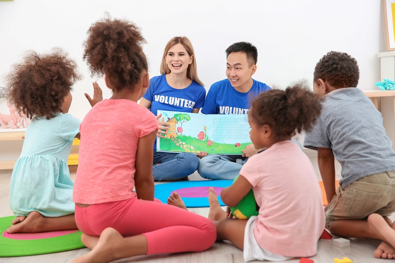 Dois voluntários, uma mulher e um rapaz, sentados em roda com quatro crianças apresentam a elas um livro infantil, em práticas de responsabilidade social.