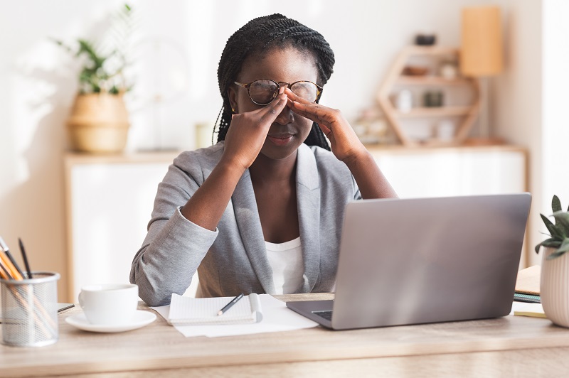 Mulher sentada à sua mesa de trabalho, com as mãos na frente do rosto e feição de cansaço, para representar uma pessoa sob estresse no trabalho.