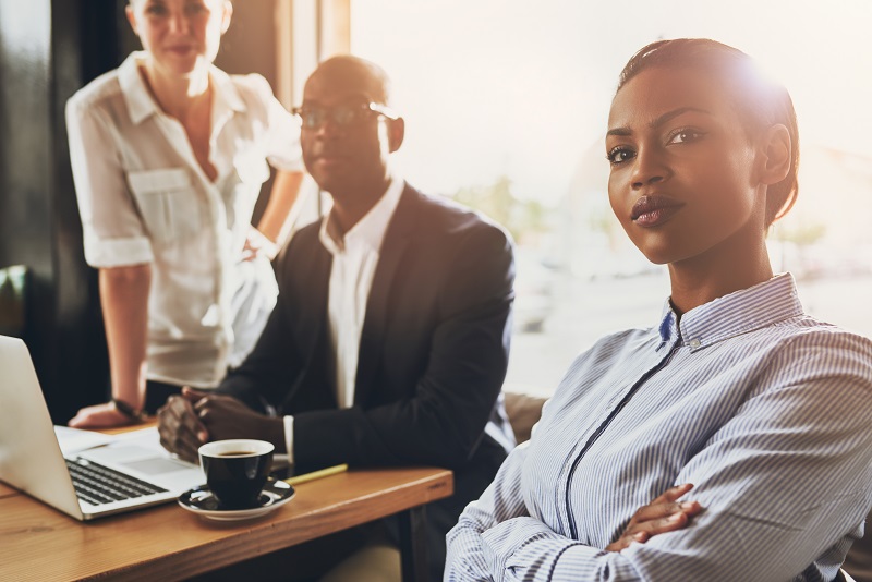 Mulher empreendedora, com roupa social, sentada a sua mesa de trabalho com sua equipe, para representar o sucesso profissional de uma pessoa com mentalidade empreendedora