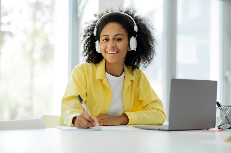 Mulher negra de cabelo solto sentada em uma mesa de escritório com seu notebook, segurando uma caneta e utilizando fones de ouvido sem fio