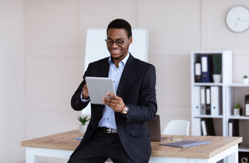 Homem negro de óculos em ambiente corporativo, vestido com roupas formais e utilizando um tablet para trabalhar