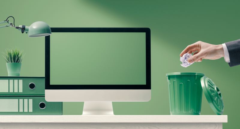 Computador sobre a mesa e uma mão jogando um papel amassado numa lixeira para papéis, simbolizando a reciclagem no trabalho.