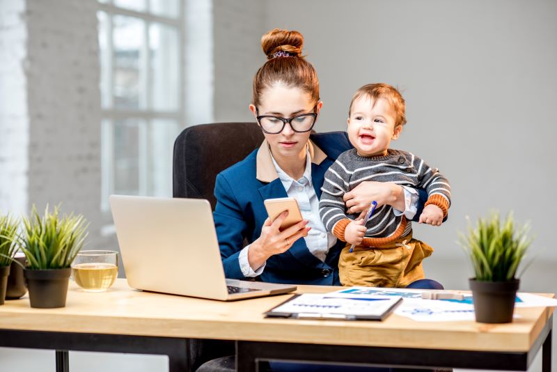 Mãe trabalhando com filho pequeno no colo enquanto mexe no celular e no computador.
