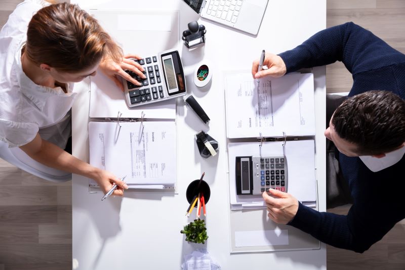 Vista de cima de dois contadores realizando contabilidade para MEI, um de frente para o outro, com papéis e calculadoras em cima da mesa.