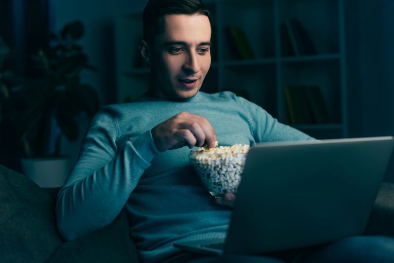 Homem assistindo filmes sobre empreendedorismo em casa, com notebook no colo enquanto come pipoca.